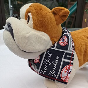 Op de afbeelding: Een witte en bruine pluche bulldog die een zwart-witte bandana draagt met de tekst "New York Yankees" en een baseballlogo. De bandana heeft rode stiksels. De bulldog zit op een zwart-witte stof met een herhalend patroon van het New York Yankees-logo, honkbalknuppels en honkballen.
