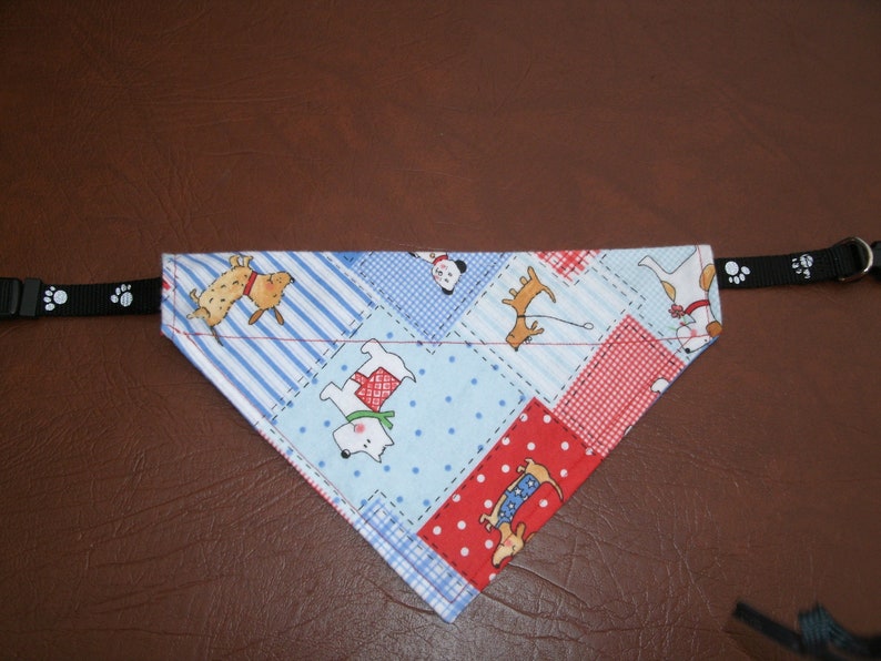 May include: A blue and red dog bandana with a patchwork design featuring various dog illustrations. The bandana is attached to a black strap with paw prints.