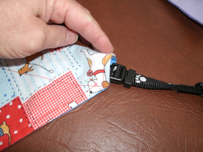 May include: A close-up of a fabric dog lead with a black plastic buckle and a paw print design. The lead is made of a patchwork fabric with a blue, white and red checked pattern.