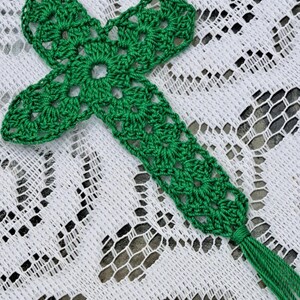 May include: A green crocheted cross bookmark with a tassel. The bookmark is on a white lace tablecloth.