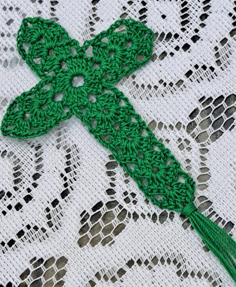 May include: A green crocheted cross bookmark with a tassel. The bookmark is on a white lace tablecloth.