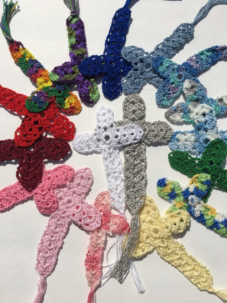 May include: A collection of handmade crocheted bookmarks in various colors and patterns, featuring a cross design. The bookmarks have tassels at the bottom.