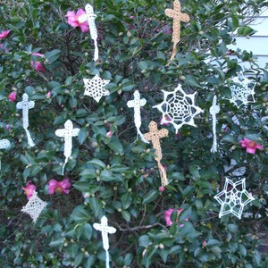 May include: A bush with pink flowers and white crocheted cross and snowflake ornaments hanging from the branches.