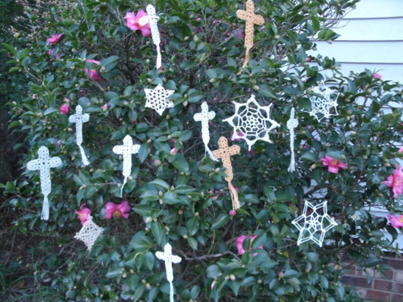 May include: A bush with pink flowers and white crocheted cross and snowflake ornaments hanging from the branches.