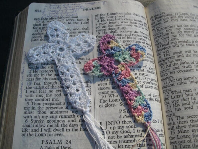 May include: A close-up of a crocheted bookmark with a floral design in shades of pink, blue, and white. The bookmark is placed on an open book of Psalms, with the text "Remember, O Lord, thy loving kindness and thy tender mercies, for they have been of old." visible.