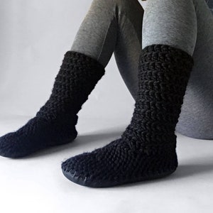May include: A pair of black knitted slipper socks with a textured pattern. The socks are worn by a person sitting on the floor.