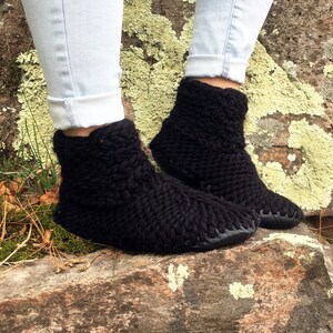May include: A pair of black knitted ankle boots with a chunky knit pattern. The boots are worn by a person wearing blue jeans and are sitting on a rock.