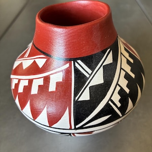 May include: A handcrafted, round ceramic vase with a wide opening. The vase features a red rim and a geometric design in red, black, and white. The design includes triangles and stepped patterns. The vase is likely used for decorative purposes.