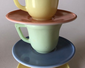 May include: A vertical stack of three ceramic cups and three saucers in pastel colors. The top cup is pale yellow, the middle cup is light green, and the bottom cup is peach. The saucers are peach, blue, and yellow, arranged in a balanced tower.