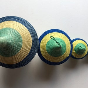 May include: A set of five conical woven baskets arranged in a descending size order. Each basket features concentric circular patterns in shades of dark blue, pale yellow, and light green, with a pointed top and a small loop at the apex.