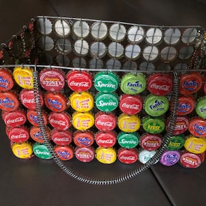 May include: A wire basket purse made from colorful bottle caps. The caps are from various brands including Coca-Cola, Sprite, Fanta, and Krest. The purse has a metal handle.