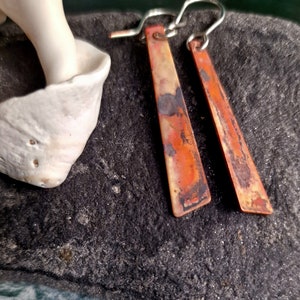 May include: A pair of long, rectangular earrings with a copper finish. The earrings have a silver hook and are made of a textured metal. The earrings are on a dark gray background.