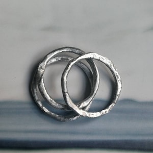 May include: Three silver rings interlocked together. The rings are made of a textured metal and have a slightly irregular shape.