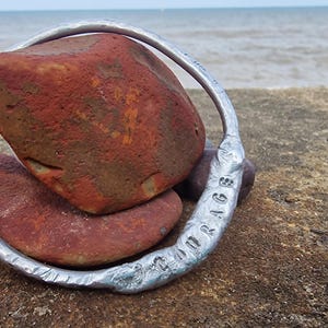 May include: A silver metal bracelet with the word "COURAGE" engraved on it, encircles two reddish-brown stones. The bracelet has a hammered texture. The background shows the ocean and a concrete surface.
