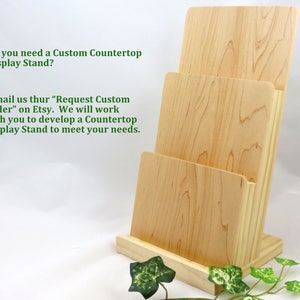 May include: A wooden countertop display stand with three tiers. The stand is made of natural wood and has a light brown color. The stand is designed to hold brochures, pamphlets, or other printed materials. The text on the stand reads: "Do you need a Custom Countertop Display Stand? E-mail us thur "Request Custom Order" on Etsy. We will work with you to develop a Countertop Display Stand to meet your needs."