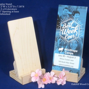May include: Unfinished wooden display stand, 8.9 cm wide, 8.3 cm deep, and 18.7 cm high, holding a brochure with the text "Best Week Ever!". The stand is designed to hold 8.9 cm x 20.3 cm literature. Pink flowers are in front of the stand.