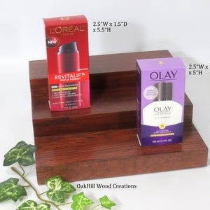 May include: Two-tiered wooden shelf with a red finish. The top shelf has a red and white bottle of L'Oreal Revitalift Triple Power Day Lotion Moisturizer with SPF 20. The bottom shelf has a purple bottle of Olay Age Defying Daily Lotion with Sunscreen SPF 15. The shelf is 2.5 inches wide, 1.5 inches deep, and 5.5 inches high. The text "OakHill Wood Creations" is printed on the bottom shelf.