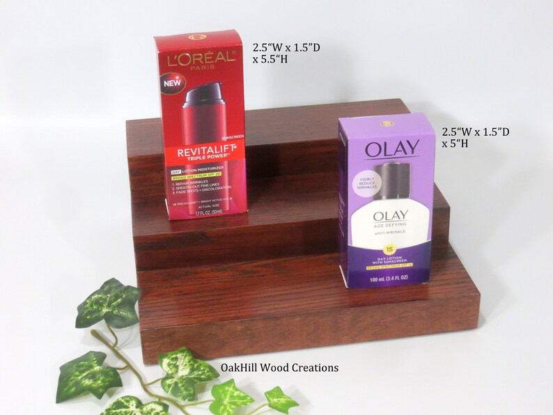 May include: Two-tiered wooden shelf with a red finish. The top shelf has a red and white bottle of L'Oreal Revitalift Triple Power Day Lotion Moisturizer with SPF 20. The bottom shelf has a purple bottle of Olay Age Defying Daily Lotion with Sunscreen SPF 15. The shelf is 2.5 inches wide, 1.5 inches deep, and 5.5 inches high. The text "OakHill Wood Creations" is printed on the bottom shelf.