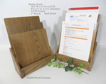 May include: A wooden display stand with three slots for holding literature. The stand is 10.5 inches wide, 6.5 inches deep, and 12.5 inches high. The slots are 8.5 inches by 11 inches or 9 inches by 12 inches. The stand has a 2-inch reveal and a husk finish. The text "OakHill Wood Creations" is visible at the bottom of the stand.