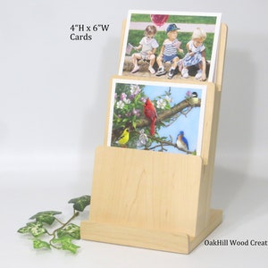 May include: A wooden card holder with two slots for displaying 4" x 6" cards. The holder is made of natural wood and has a simple, elegant design. The top slot displays a card with a picture of three children, while the bottom slot displays a card with a picture of birds on a branch.
