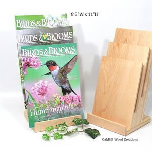 May include: A wooden magazine rack with three slots to hold magazines. The rack is made of natural wood and has a simple design. The rack is 8.5 inches wide and 11 inches high. Three copies of Birds & Blooms magazine are displayed in the rack. The magazine cover features a hummingbird in flight.