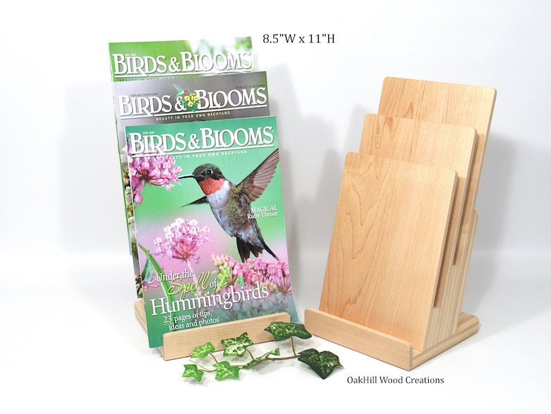 May include: A wooden magazine rack with three slots to hold magazines. The rack is made of natural wood and has a simple design. The rack is 8.5 inches wide and 11 inches high. Three copies of Birds & Blooms magazine are displayed in the rack. The magazine cover features a hummingbird in flight.