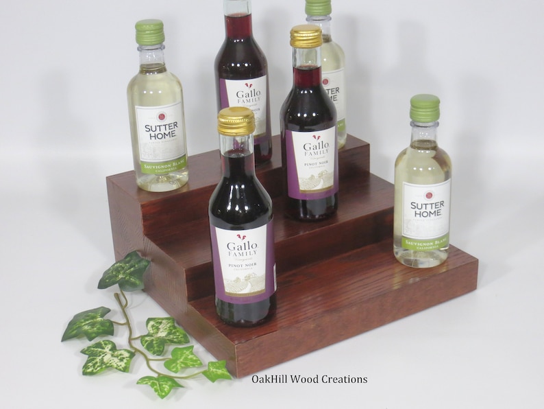 May include: A three-tiered wooden wine rack with a dark brown stain. The rack is filled with five miniature bottles of wine, including Sutter Home Sauvignon Blanc and Gallo Family Pinot Noir.