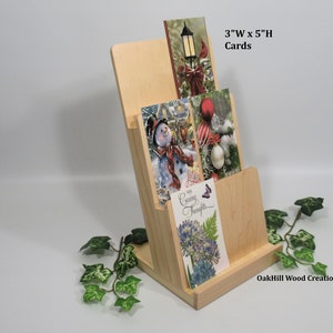 May include: A wooden card holder with three slots for displaying greeting cards. The holder is made of natural wood and has a simple, elegant design. The cards are 3 inches wide by 5 inches high. The holder is labeled "OakHill Wood Creations".