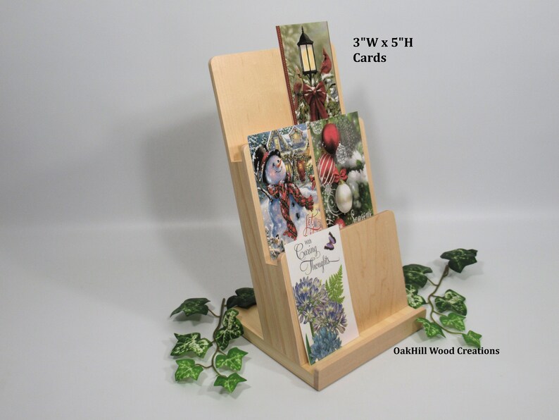 May include: A wooden card holder with three slots for displaying greeting cards. The holder is made of natural wood and has a simple, elegant design. The cards are 3 inches wide by 5 inches high. The holder is labeled "OakHill Wood Creations".