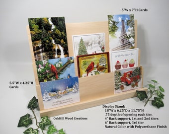 May include: A natural wood card display stand with three tiers. The stand is 18 inches wide, 6.25 inches deep, and 11.75 inches high. The stand has a 0.75 inch depth opening for each tier. The stand has a 4 inch back support for the first and second tiers and a 6 inch back support for the third tier. The stand is made of natural wood with a polyurethane finish. The stand is holding a variety of cards, including Christmas cards, birthday cards, and other greeting cards.