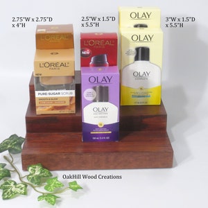 May include: Three-tiered wooden display stand with four bottles of skincare products. The top tier has two bottles of L'Oreal Paris Pure Sugar Scrub, the middle tier has a bottle of Olay Age Defying Day Lotion, and the bottom tier has a bottle of Olay Complete Daily Moisturizer. The stand is made by OakHill Wood Creations.