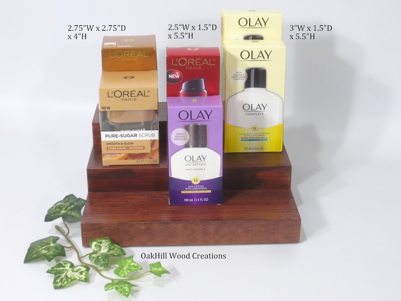 May include: Three-tiered wooden display stand with four bottles of skincare products. The top tier has two bottles of L'Oreal Paris Pure Sugar Scrub, the middle tier has a bottle of Olay Age Defying Day Lotion, and the bottom tier has a bottle of Olay Complete Daily Moisturizer. The stand is made by OakHill Wood Creations.