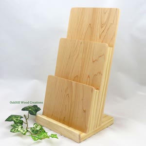 May include: A wooden magazine rack with three slots, made of light wood with a natural finish. The rack is standing on a white surface. OakHill Wood Creations is printed on the side of the rack.