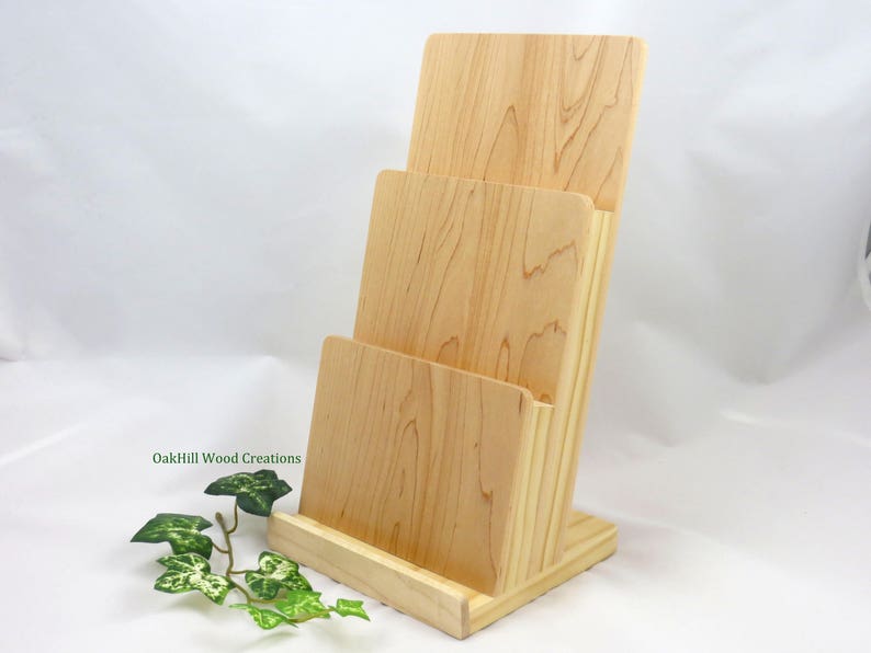 May include: A wooden magazine rack with three slots, made of light wood with a natural finish. The rack is standing on a white surface. OakHill Wood Creations is printed on the side of the rack.