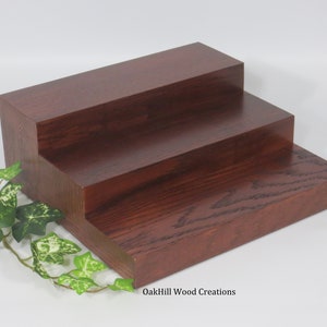 May include: A three-tiered wooden display stand made of oak. The stand is stained a rich mahogany color and has a natural wood grain finish. The stand is perfect for displaying small items, such as jewelry, candles, or plants. OakHill Wood Creations.