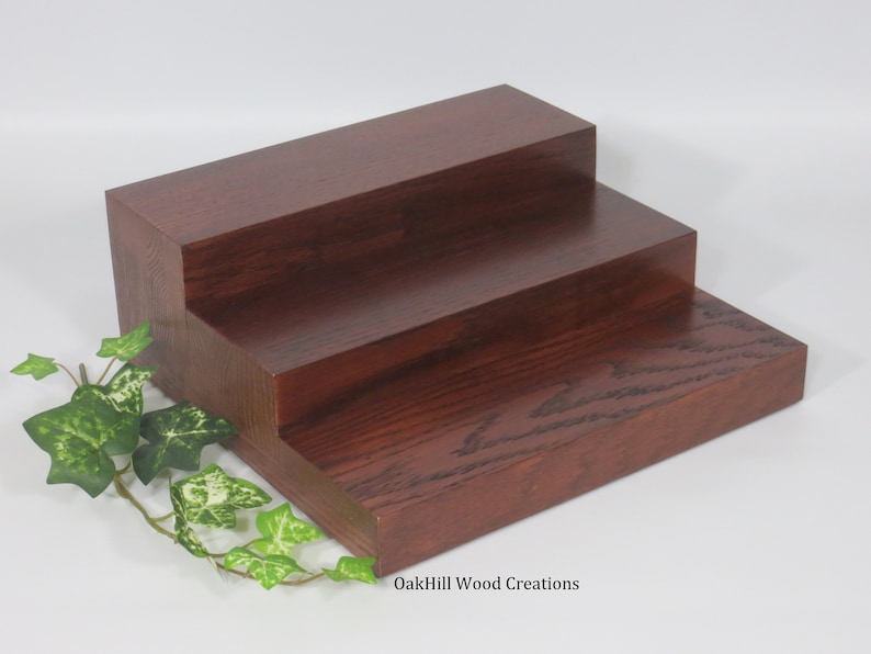 May include: A three-tiered wooden display stand made of oak. The stand is stained a rich mahogany color and has a natural wood grain finish. The stand is perfect for displaying small items, such as jewelry, candles, or plants. OakHill Wood Creations.