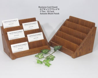 May include: Two wooden business card stands with five tiers, each holding business cards. The stands are made of a dark brown wood with a slightly reddish hue and have a natural wood grain finish. The stands are 8.5 inches wide, 5.75 inches deep, and 6 inches high. The stands are labeled "OakHill Wood Creations" and "Autumn Blush Finish".