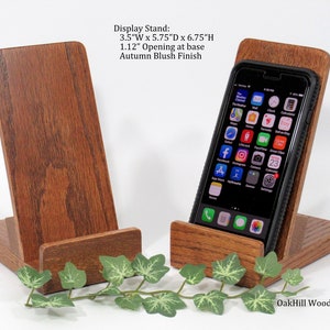 May include: Two wooden phone stands in an autumn blush finish. The stand on the right has a black smartphone in it. The stands are 3.5 inches wide, 5.75 inches deep, and 6.75 inches high. The opening at the base of the stand is 1.12 inches.