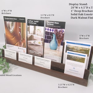 May include: A dark walnut wood brochure stand with five slots for brochures. The stand is 20 inches wide, 4.5 inches deep, and 5 inches high. The stand has a 1 inch deep opening for brochures. The stand is made of solid oak and has a dark walnut finish. The stand is holding brochures of various sizes, including a 6 inch by 9 inch brochure, a 3.5 inch by 8.5 inch brochure, a 5 inch by 5 inch brochure, and a 3.25 inch by 4.25 inch brochure.