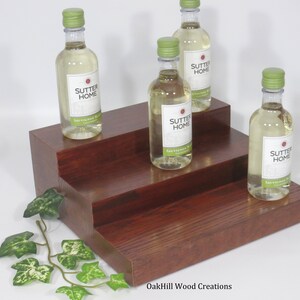 May include: A three-tiered wooden wine bottle display stand with a dark reddish-brown finish. Four bottles of Sutter Home wine are displayed on the stand. The stand is made by OakHill Wood Creations.
