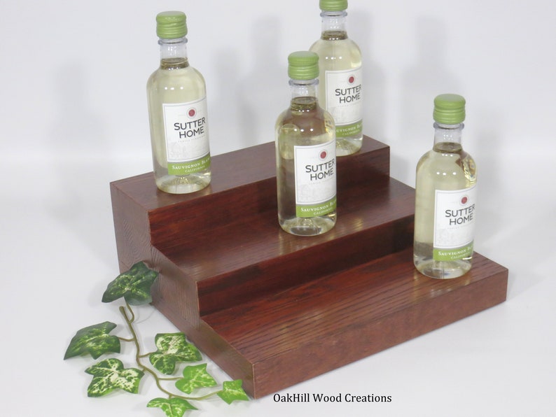 May include: A three-tiered wooden wine bottle display stand with a dark reddish-brown finish. Four bottles of Sutter Home wine are displayed on the stand. The stand is made by OakHill Wood Creations.