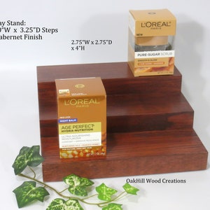 May include: A three-tiered wooden display stand with a Cabernet finish. The stand is 10 inches wide, 3.25 inches deep, and 4 inches high. The stand is displaying two L'Oreal Paris skincare products: a jar of Age Perfect Hydra-Nutrition Night Balm and a jar of Pure-Sugar Scrub. The stand is made by OakHill Wood Creations.