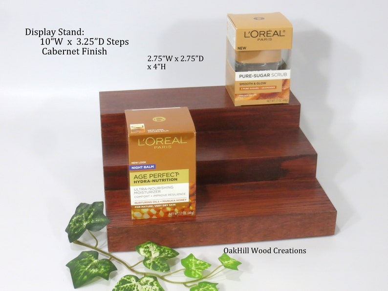May include: A three-tiered wooden display stand with a Cabernet finish. The stand is 10 inches wide, 3.25 inches deep, and 4 inches high. The stand is displaying two L'Oreal Paris skincare products: a jar of Age Perfect Hydra-Nutrition Night Balm and a jar of Pure-Sugar Scrub. The stand is made by OakHill Wood Creations.