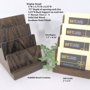 May include: A dark brown wooden gift card display stand with four tiers. The stand is 5 inches wide, 5.75 inches deep, and 6.25 inches high. The stand has a Jacobean stain finish. The stand is holding four gift cards that say "Gift Card" in black text on a light yellow background. The gift cards are 2 inches wide and 3.5 inches high. The stand is also designed to hold envelopes that are 2.5 inches wide and 4.56 inches high.