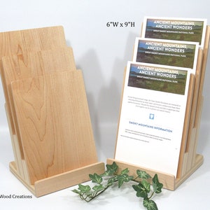 May include: A wooden brochure holder with a natural finish. The holder is 15.2 cm wide and 22.9 cm tall. It has four slots for brochures and a base that is angled to hold the brochures upright. The holder is made of maple wood and has a natural wood grain. The brochures in the holder are for the Great Smoky Mountains National Park and feature the text "Ancient Mountains, Ancient Wonders".