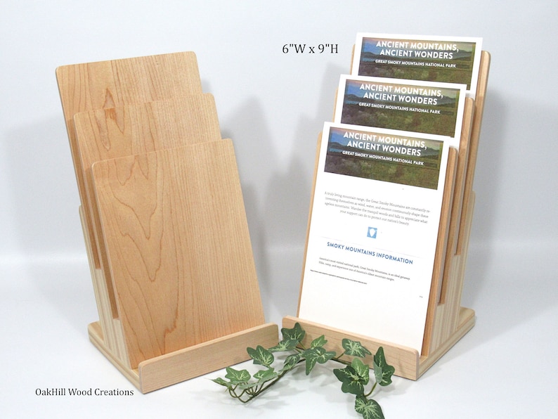 May include: A wooden brochure holder with a natural finish. The holder is 15.2 cm wide and 22.9 cm tall. It has four slots for brochures and a base that is angled to hold the brochures upright. The holder is made of maple wood and has a natural wood grain. The brochures in the holder are for the Great Smoky Mountains National Park and feature the text "Ancient Mountains, Ancient Wonders".