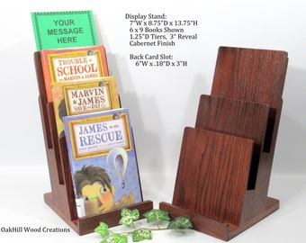 May include: A wooden book display stand with three tiers, each tier is 1.25 inches deep and can hold a book up to 6 inches wide by 9 inches tall. The stand is 7 inches wide by 8.75 inches deep by 13.75 inches tall. The stand has a back card slot that is 6 inches wide by 0.18 inches deep by 3 inches tall. The stand is made of wood and has a dark red finish. The stand is shown with books on it. The books are titled "Trouble at School", "Marvin & James Save the Day", and "James to the Rescue". The stand is made by OakHill Wood Creations.