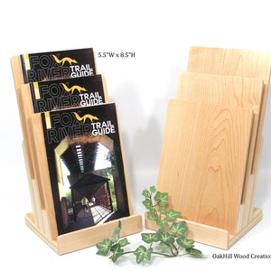 May include: A wooden magazine rack with three slots for holding brochures or pamphlets. The rack is made of natural maple wood and measures 14 cm wide by 21.5 cm high. The rack is displayed with brochures that say "Fox River Trail Guide".
