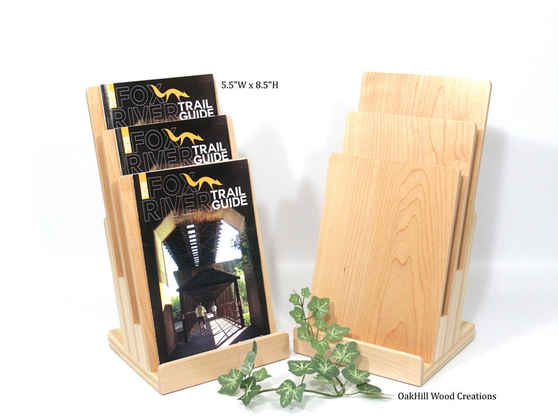 May include: A wooden magazine rack with three slots for holding brochures or pamphlets. The rack is made of natural maple wood and measures 14 cm wide by 21.5 cm high. The rack is displayed with brochures that say "Fox River Trail Guide".