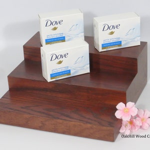 May include: Three Dove gentle exfoliating body wash bars displayed on a three-tiered wooden stand. The stand is made of dark wood with a reddish brown hue. The stand is on a white surface. The Dove bars are white with blue and gold accents. The text on the bars reads "Dove gentle exfoliating exfoliant douceur beauty bar - pain de beaut&eacute;"